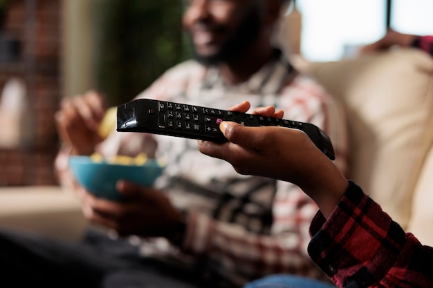 Girlfriend holding tv remote control to switch channel program and find movie on television to watch. Eating takeaway delivery from fast food and watching film together. Close up.