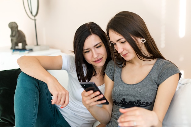 The girls are sitting on the couch chatting and looking at the phone. 