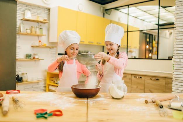 Girls cooks in caps, cookies preparation on the kitchen. Kids cooking pastry, little chefs makes dough, child preparing cake