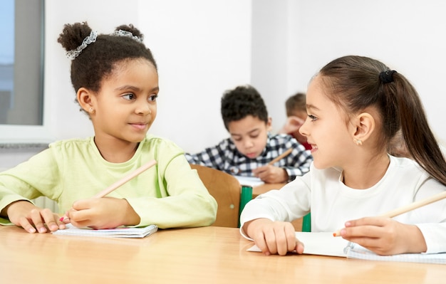 Photo girls at desk in classroom, looking at each other, posing.