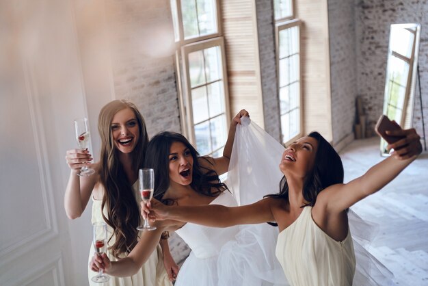 Photo girls going crazy. top view of attractive young bride and two bridesmaids making faces while taking selfie in the fitting room