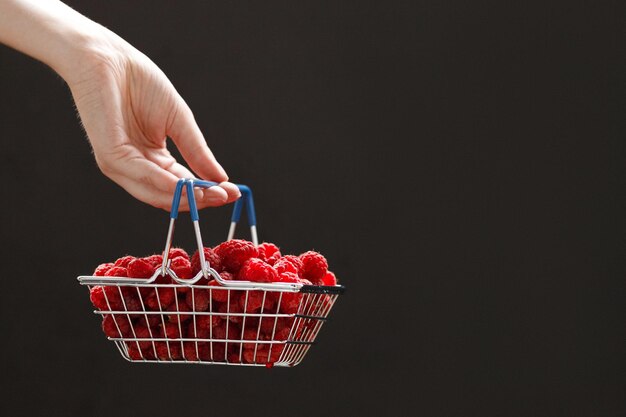 Girls hand holding a mini grocery shopping basket with fresh raspberries on a dark background