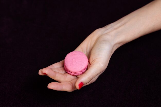 Photo girls hand with bright pink manicure hold bright pink halves of macarons on dark background closeup view making dessert