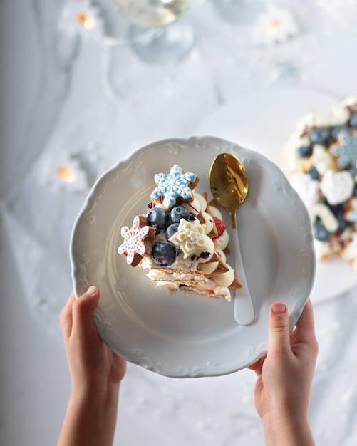 Girls hands holding white plate with piece of christmas cake decorated whipped cream gingerbread cookies blueberry Vertical