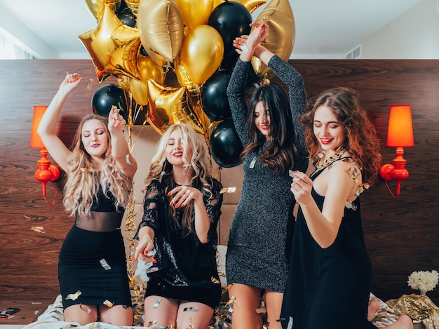 Girls hangout fun. Great time together. Joyful young females dancing on bed. Bachelorette party. Confetti and balloons.