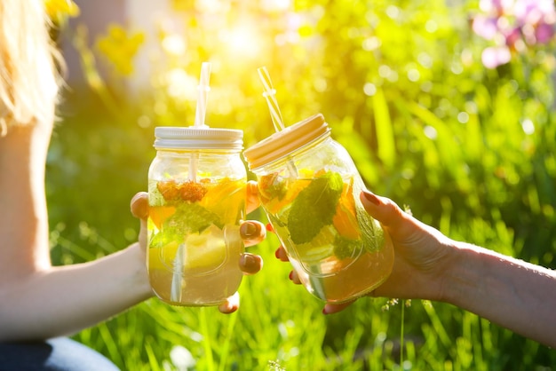 ストローで瓶に新鮮なレモネードを保持している女の子。流行に敏感な夏の飲み物。自然の中で環境にやさしい。ガラスにミントが入ったレモン、オレンジ、ベリー。屋外の緑の高い草。