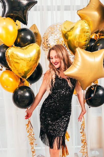 Girls party. Special occasion. Beautiful lady in black sparkling dress smiling, standing with balloons over white curtains.