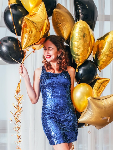 女の子のパーティー特別な機会白いカーテンの上に風船で立って笑顔の青い輝くドレスで幸せな陽気な女性