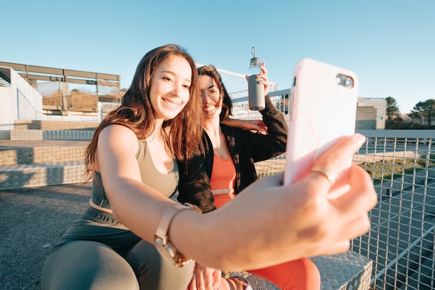 Girls taking a selfie while resting from work out sitting on stairs, modern sporty youngsters recording a video for social networks while smiling. Good vibes and happiness concept. African girls