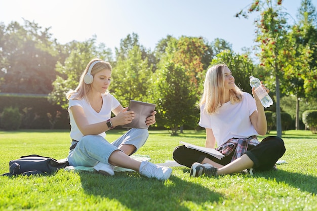 Girls teenage students sitting on green lawn in park with backpack, books, digital tablet, drink bottle of water. University, college, school, education and knowledge, adolescent lifestyle