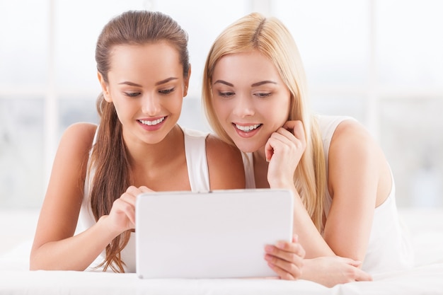 Girls with digital tablet. Two attractive young women lying on bed together and using digital tablet