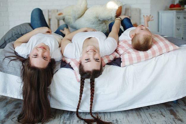 Girls with pigtails and a younger brother, a family in white T-shirts and jeans play with pillows on the bed. The concept of a happy family.