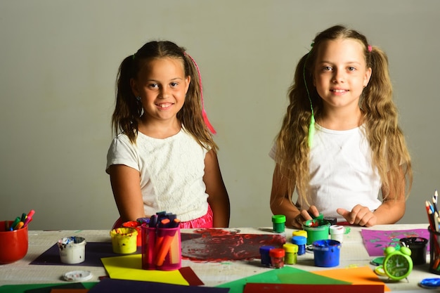 Girls with smiling faces by their art desk Children paint with gouache and stationery on grey background Artists with ponytails create messy artwork Creativity and imagination concept