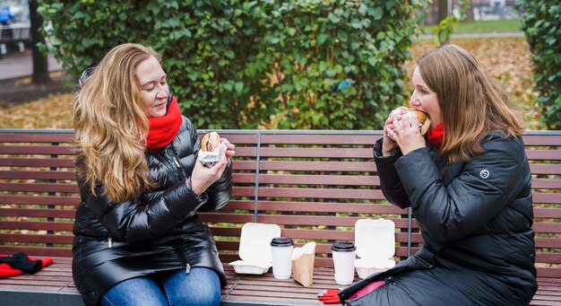 Girlswomen friends meetingeating hamburgercheeseburgerJunk fatty tasty street foodTake away burgerLunch breakweekend outdoors in park sitting on benchUnhealthy dietBody positiveoverweight