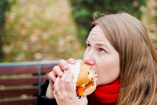 Girlwoman Eating big hamburgercheeseburgerジャンク 脂肪の多いおいしい屋台の食べ物ハンバーガーを持ち帰りベンチに座って公園で屋外で昼休み不健康な食事週末のリラックスボディポジティブ太りすぎの問題