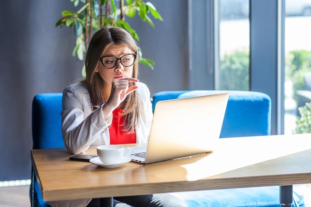 Give me a few more. Portrait of pleased stylish young woman in glasses sitting, looking at her laptop screen on video call and asking for something more. indoor studio shot, cafe, office background.