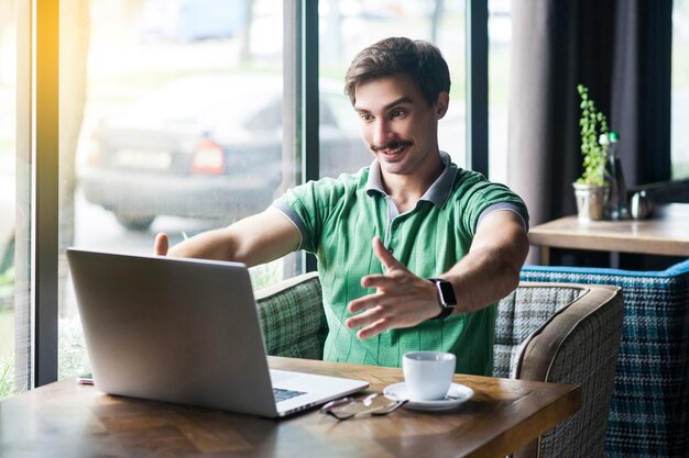 Give me hug Young happy businessman in green tshirt sitting and looking at laptop on video call and want to embrace and toothy smile business concept indoor shot near big window at daytime