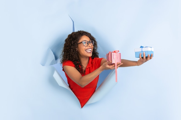 Giving presents, gifts. Cheerful african-american young woman in torn blue paper background, emotional, expressive. Breaking on, breakthrought. Concept of human emotions, facial expression, sales, ad.