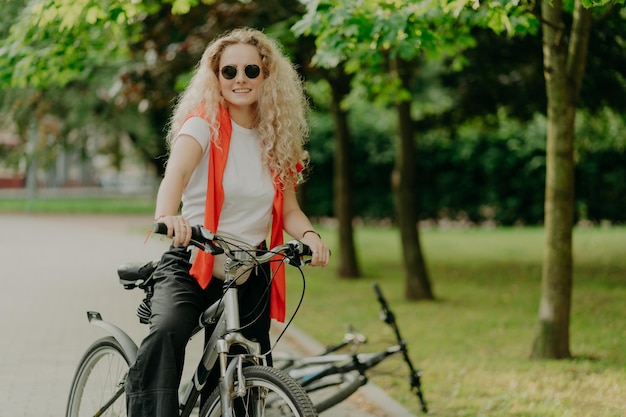 Glad active curly haired female bicyclist covers distance on
her own transport wears sunglasses casual outfit poses in park with
green trees expresses good emotions riding bicycle concept