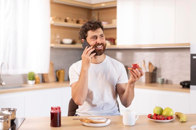 Glad adult caucasian guy with beard in white tshirt eats sandwich calls by phone in modern kitchen interior