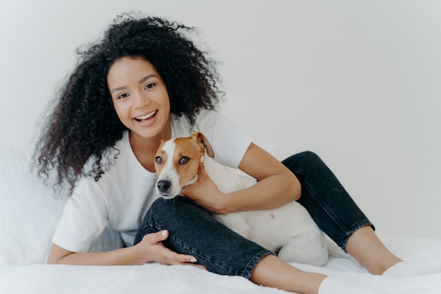 Glad afro woman rests in bed with dog have playful mood pose together in bedroom against white background