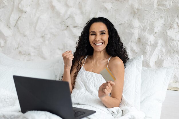 Glad arab millennial curly lady woke up sitting on white bed with laptop making victory and success gesture