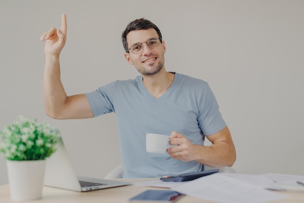 Glad attractive brunet young male in round glasses drinks tea or coffee sits at working place works with modern technologies raises hand as gets good idea for developing new financial project