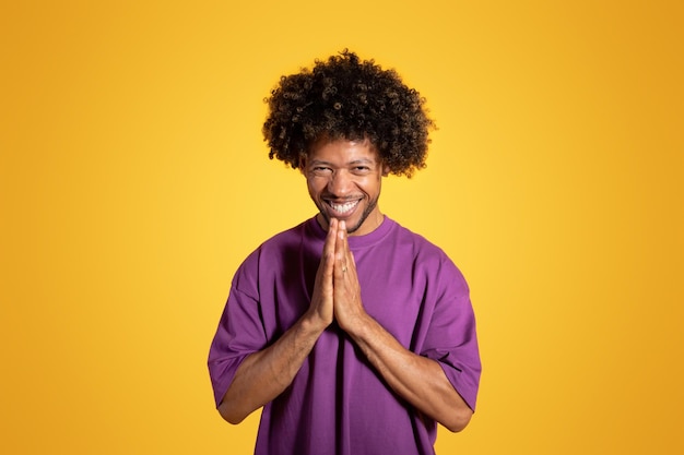 Glad black middle aged curly man in violet tshirt prays with hands isolated on orange background
