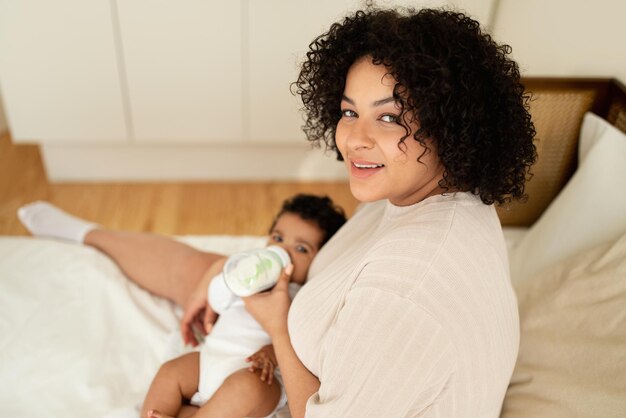 Photo glad black millennial mom feeds with bottle small baby on white bed enjoys kid care in bedroom