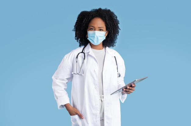 Glad black millennial woman doctor in white coat and protective mask with tablet enjoy work exam