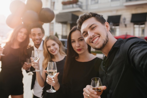 Glad caucasian man with dark hair making selfie on the street with friends, carrying black party balloons. Outdoor photo of smiling guy in trendy shirt taking picture with colleague during travel.