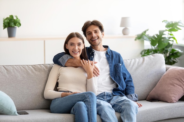 Glad cheerful millennial caucasian guy hugs woman on sofa and looking at camera in living room