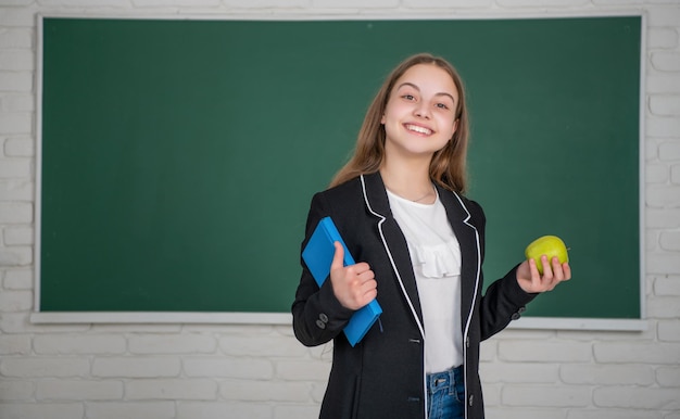 Glad child standing on blackboard background with workbook and apple at school