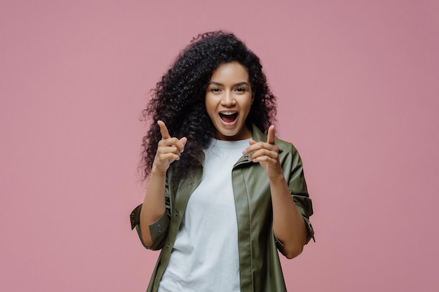 Glad confident woman with afro hair points index fingers\
directly at camera chooses you picks someone in her team dressed in\
white t shirt and leather shirt poses against pink background