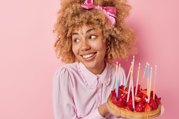 Glad curly haired woman poses with delicious cake looks away
with positive smile on face dressed in festive clothes celebrates
birthday stands against pink background blank space for your
text