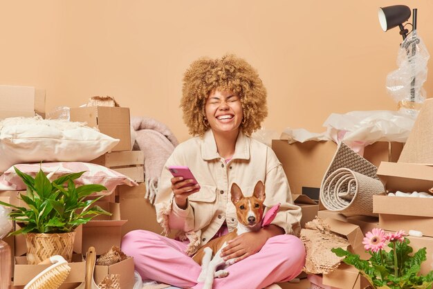 Glad curly haired woman smiles toothily holds mobile phone\
searches new apartment for renting sits crossed legs with pet\
dressed in casual clothes collects pesonal belongings in carton\
boxes