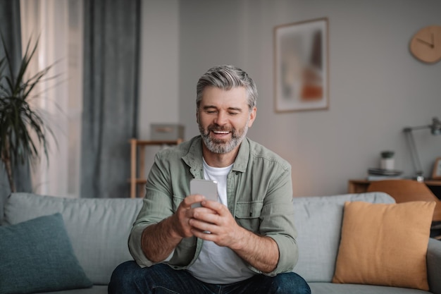 Glad elderly european man with beard chatting on smartphone watching video playing game