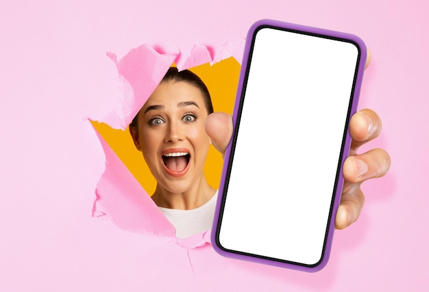 Glad excited shocked young caucasian woman looking through hole in pink paper and showing smartphone