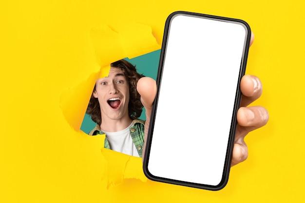 Glad excited young caucasian guy with open mouth looks through hole in yellow paper and shows