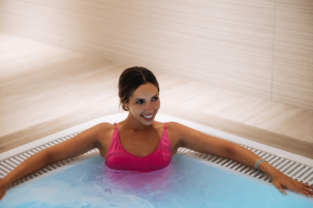수영복을 입은 행복한 여성은 온수 욕조에 앉아 거품을 일으키며 주말에 스파 절차를 하는 동안 휴식을 취합니다.