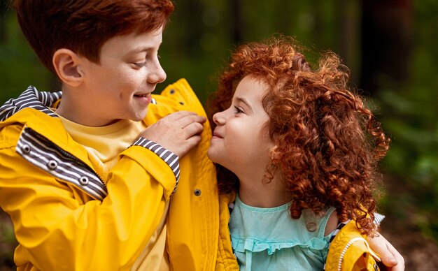 Glad ginger boy in yellow raincoat smiling and embracing sister with curly hair on weekend day in countryside