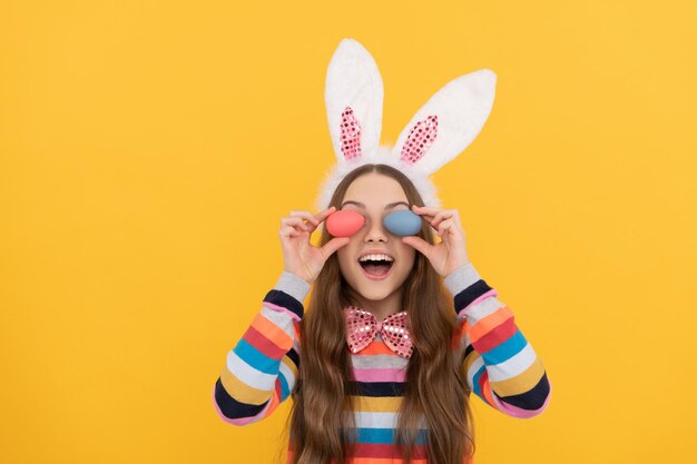 Glad girl in bunny ears and bow tie hold easter eggs on yellow background