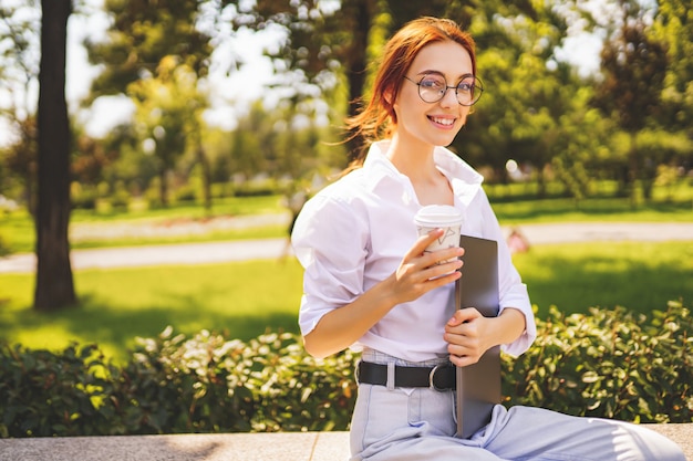 Glad girl student in glasses and white shirt holds laptop and coffee to go while sitting in the park