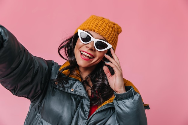 Foto ragazza felice con occhiali da sole e cappello arancione che si fa selfie studio shot di donna sorridente in piumino isolato su sfondo rosa