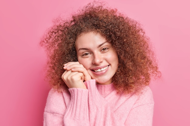 Glad good looking young woman with curly bushy hair keeps hands near face expresses positive emotions expresses sincere feelings poses against pink  wall. Pleasant emotions concept