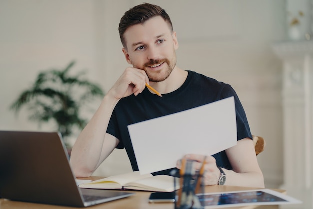 Glad handsome European businessman holds paper document poses in home interior at desktop surrounded by modern technologies checks information has remote work looks thoughtfully into distance