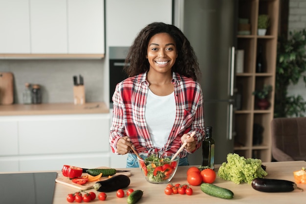 Glad happy pretty black young female chef in casual prepares salad from organic vegetables