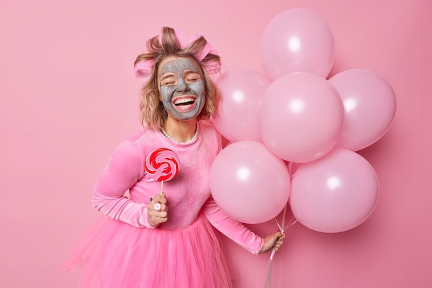 Glad happy woman applies beauty clay mask on face giggles positively holds sweet candy on stick and bunch of helium balloons wears festive dress makes hairstyle with hair rollers enjoys celebration