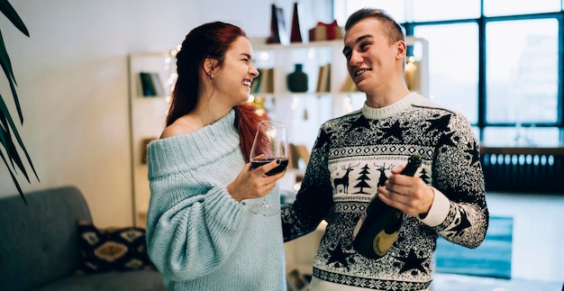 Glad laughing couple in cozy wear with glass and bottle of wine cuddling and celebrating Christmas