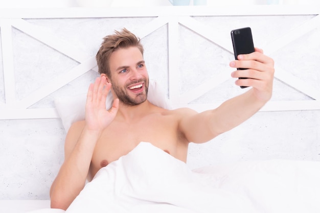 Радостный мужчина делает селфи на телефоне и машет приветствием в белой спальне